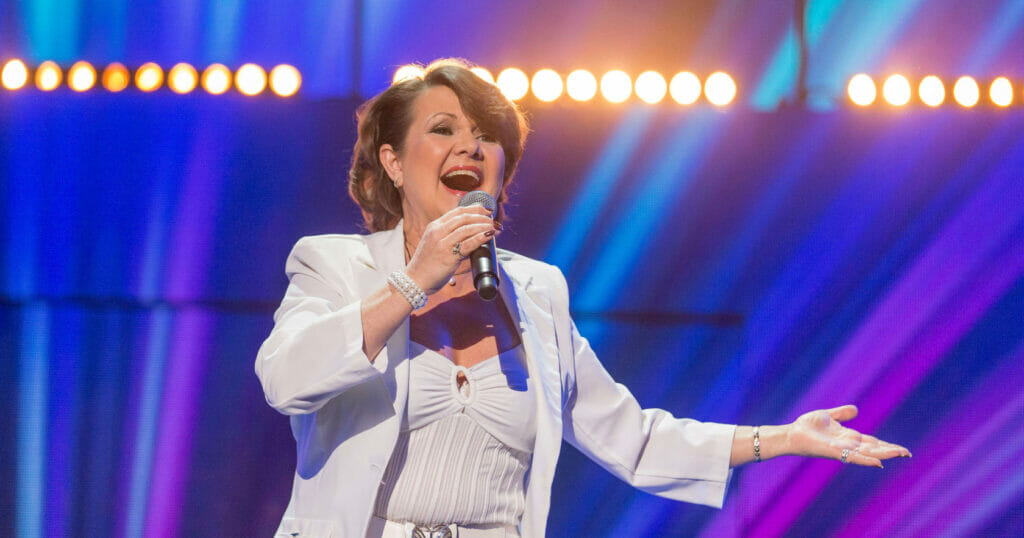 A woman in a white suit singing into a microphone at a concert.