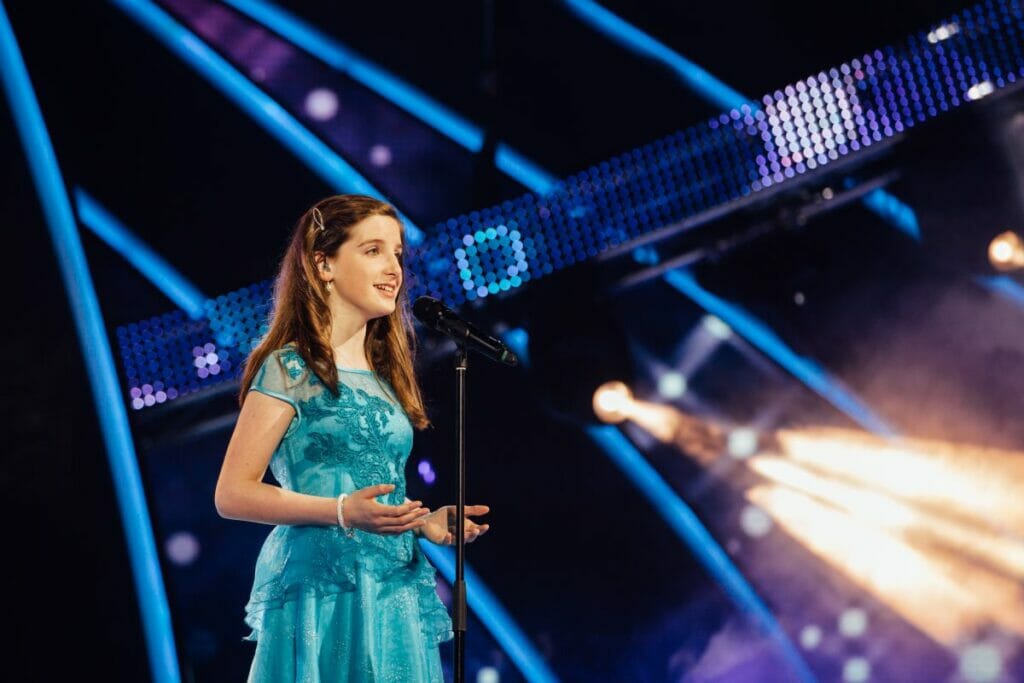 A young girl in a blue dress singing into a microphone with grace and passion.