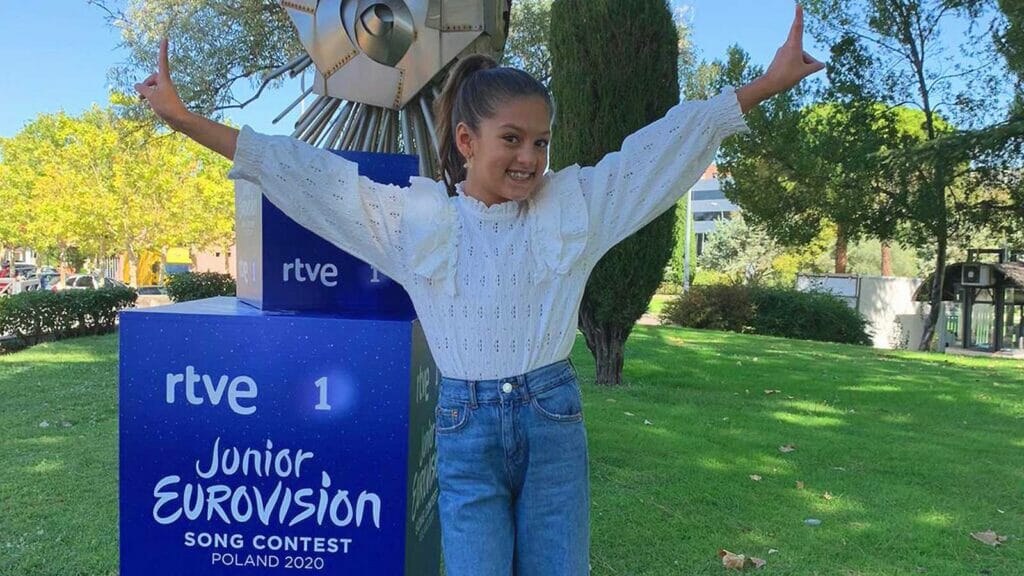 A girl posing in front of a sign that says "rive junior eurovision".