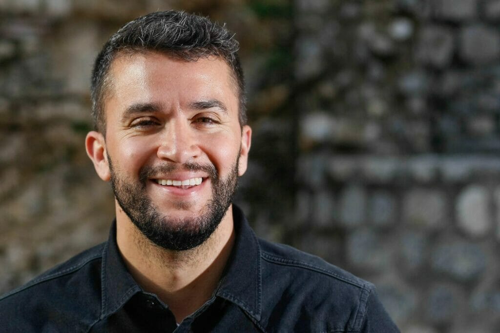 A bearded man grinning in front of a stone wall.