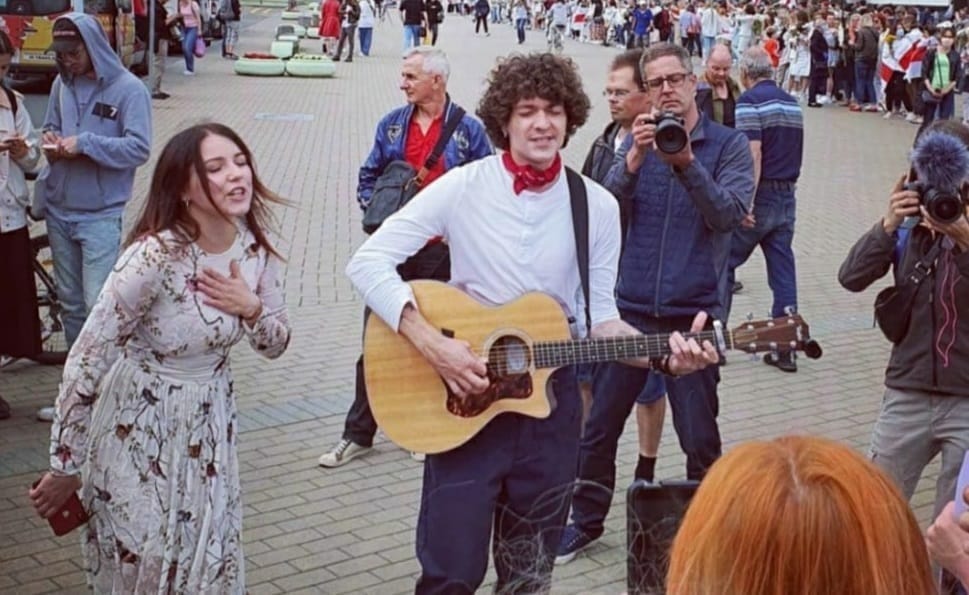 A man and a woman singing and playing a guitar in a crowded environment.